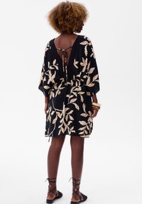 Zwarte jurk met gouden bladrandpatroon, korte mouwen, open rug geknoopt met een lint, cinched taille en zijsplitten, gedragen met zwarte gespsandalen.