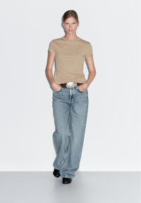 Béžové přiléhavé tričko v kombinaci s vysoce pasovými světle modrými širokými džínami, doplněné hnědým páskem se stříbrnou sponou a černými botami.