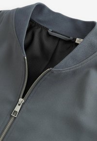 Veste grise avec fermeture éclair argentée partiellement ouverte, montrant une doublure en mesh noire et une étiquette de taille à l'intérieur du col.