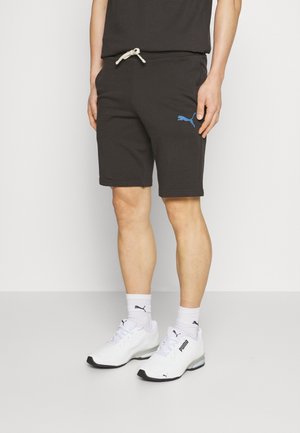 Shorts noirs en mélange de coton, avec une taille à cordon de serrage et des poches latérales. Un logo Puma bleu est présent sur la cuisse gauche. Porté avec des baskets blanches.