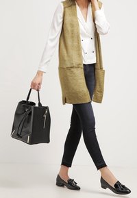 Lång, öppen olivväst, vit blus med dubbelknappstängning, mörka jeans, svart strukturerad väska och svarta loafers med rosetter.