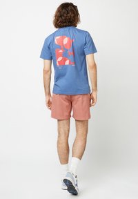 T-shirt in cotone blu con design grafico astratto rosso e rosa sulla schiena, abbinato a pantaloni corti rosa chiaro e sneaker blu.