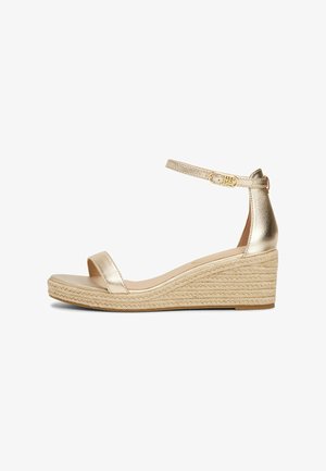 Sandale compensée métallique dorée avec une semelle en jute tissée. Elle dispose d'une bride de cheville avec une boucle et d'un design à orteils ouverts, avec une texture lisse et des accents minimaux.