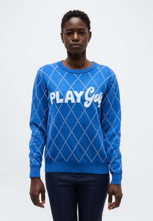 Henkilö pukeutuneena siniseen argyle-neuleeseen, jossa lukee "PLAY Golf", seisoo yksinkertaisen vaalean taustan edessä katsellen eteenpäin.