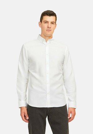 Camisa de algodón blanca de manga larga con cuello american, botones delanteros y una textura suave. Lleva puestos unos pantalones de mezclilla oscuros.