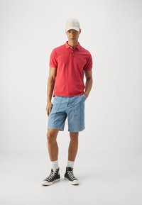 Punase polo särk, heledad sinised denim lühikesed püksid taskutega, mustad kõrged tennisetossud ja beež pesapalli müts. Ühetooniline taust.