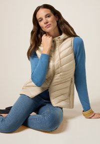 Beige gesteppte Weste mit Kapuze, Reißverschluss vorne und seitlichen Taschen, getragen über einem blauen Pullover und blauen Jeans. Glatte Textur und taillierte Form.