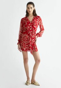 Red floral dress with sheer fabric, ruffle detail at the neckline, long sleeves, cinched waist, and a flowy hem. Paired with gold slip-on shoes.