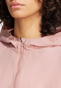 Ljusrosa zip-jacka med huva, texturerad tyg och central dragkedja. Närbild som lyfter fram detaljerna på kragen och dragkedjan.