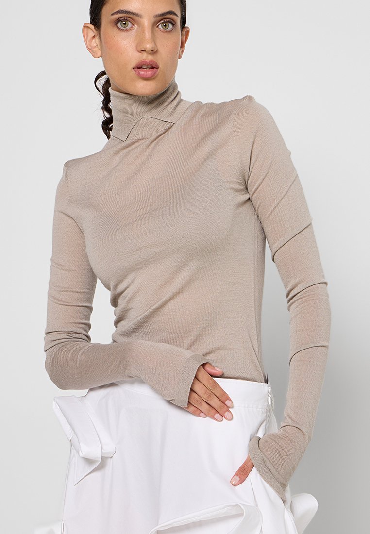 Beige Rollkragenoberteil mit langen Ärmeln und Daumenlöchern, aus einem leichten, gerippten Stoff, kombiniert mit einem weißen Rock mit Rüschendetails.