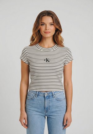 Mujer joven con cabello ondulado que lleva un top corto a rayas negras y blancas y unos vaqueros de talle alto azul claro sobre un fondo liso.