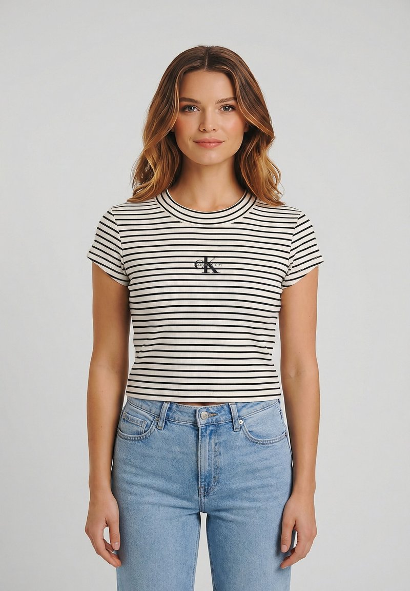 Jeune femme aux cheveux ondulés portant un crop top rayé noir et blanc et un jean taille haute bleu clair, sur un fond uni.
