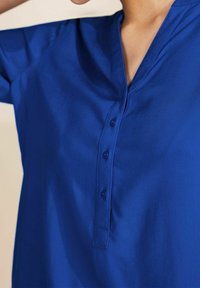 Camicia azzurra con colletto a V, maniche corte e tessuto morbido. Ha quattro bottoni sulla parte anteriore per la chiusura.