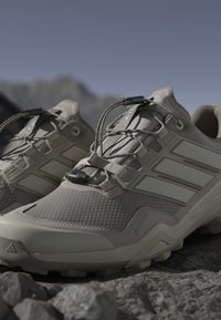 Beige outdoor shoes with a textured upper, lacing system, and patterned sole. Features branded accents and reinforced toe for durability.