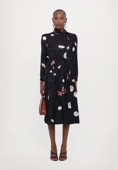 Zwarte bloemen midi-jurk met lange mouwen, strikdetail bij de hals en roze en witte bloemmotieven. Model houdt een bruine handtas vast en draagt zwarte sandalen.