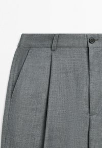 Szare spodnie na miarę z przednią zakładką, boczną kieszenią, szlufką na pasek i czarnym zapięciem na guzik na białym tle.