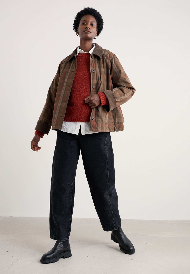 Brūns un svītrainas pogājamā jaka ar korduroja apkakli, kas uzlikta pār sarkano džemperi un baltu krekli, kopā ar melnu platu apakšbiksēm un melnu apaviem.