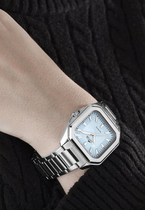 Srebrna ura iz nerjavečega jekla s kvadratnim številčnico, svetlo modrim številčnico, srebrnimi označevalci ur in teksturiranim črnim puloverjem iz pletenega kabla v ozadju.