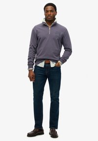 Grijze zip-up sweatshirt met logo, laagjes over een lichtblauw gestreept shirt. Donkerblauwe jeans en bruine instappers maken de outfit compleet.