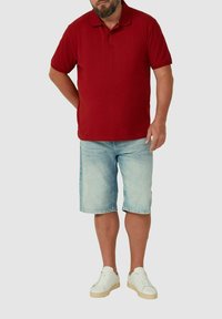 Mann steht und trägt ein rotes Polohemd, helle blaue Jeansshorts und weiße Sneaker vor einem einfarbigen Hintergrund.