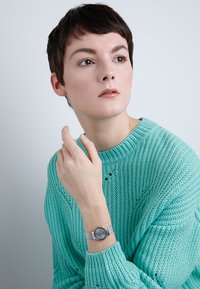 Srebrny siateczkowy zegarek na nadgarstek z okrągłą tarczą, charakteryzujący się minimalistycznym designem i subtelnymi znacznikami godzin, noszony na tle teksturowanego turkusowego swetra.