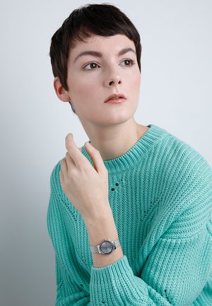 Silbernes Netzarmbanduhr mit rundem Zifferblatt, das ein minimalistisches Design und subtile Stundenmarkierungen aufweist, getragen über einem strukturierten türkisfarbenen Strickpullover.