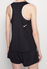 Zwart atletisch tanktop met een open rug en een witte Nike-logo. Het materiaal lijkt licht en ademend, gecombineerd met bijpassende shorts.