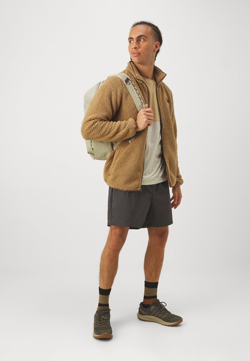 Giacca in pile marrone, t-shirt beige, pantaloni corti scuri e scarpe da trekking grigie. Presenta uno zaino leggero e calze a righe. Insieme orientato al fitness.