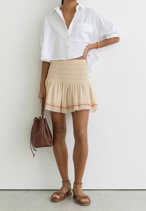 Person iført en hvid cropped skjorte, beige smocket mininederdel med røde striber, brune sandaler og holder en brun læder bucket taske.