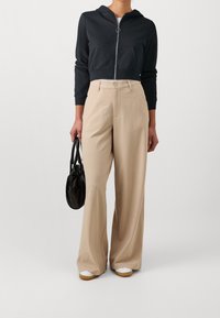 Mujer vistiendo pantalones de pierna ancha beige, sudadera negra con cremallera corta, zapatillas blancas y sosteniendo un bolso negro frente a un fondo sencillo.