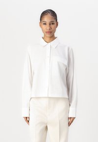 Hvit cropped skjorte med knapp, krage, lange ermer og en enkelt brystlomme; har en glatt tekstur og et enkelt design.