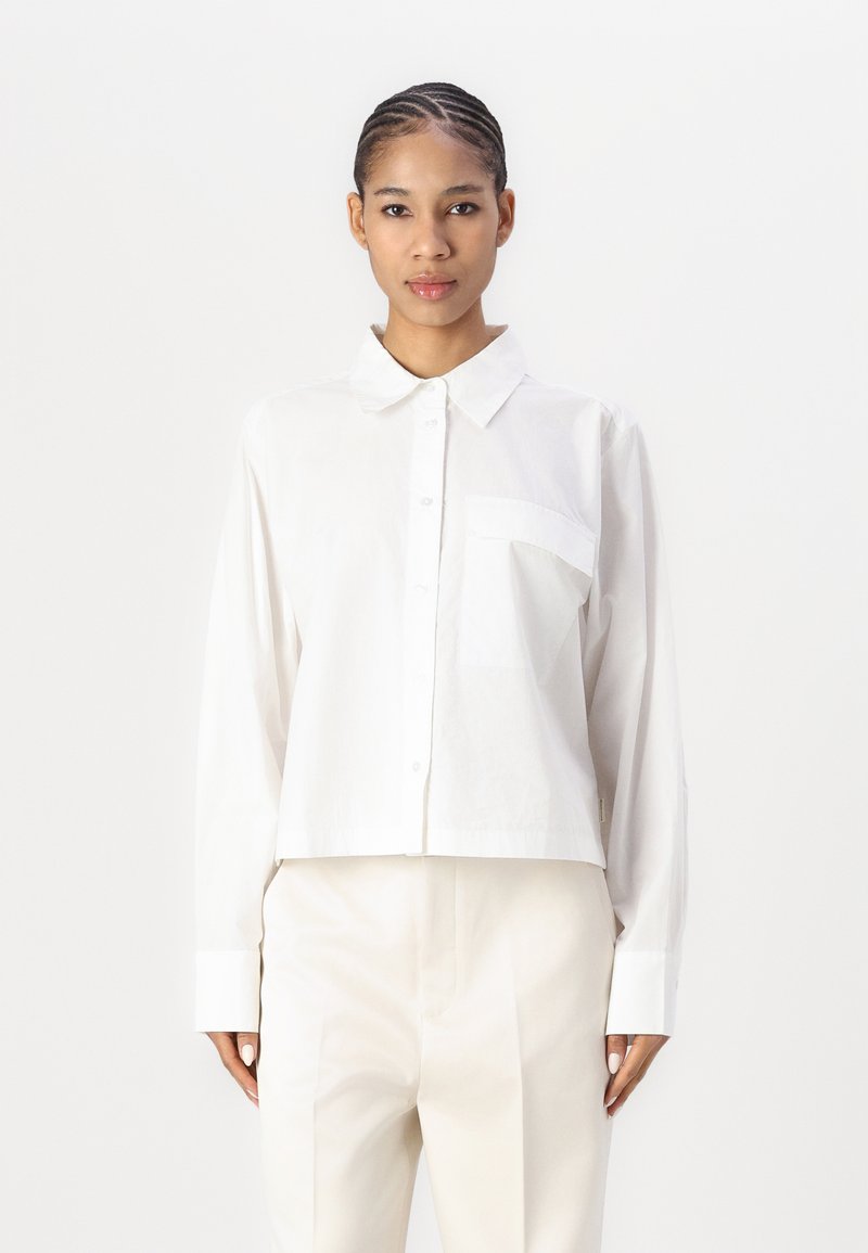 Hvit cropped skjorte med knapp, krage, lange ermer og en enkelt brystlomme; har en glatt tekstur og et enkelt design.