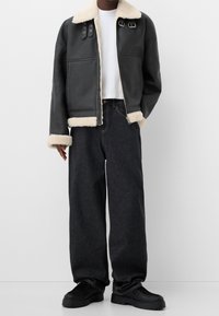 Personne portant une veste en shearling noire avec doublure crème, un t-shirt blanc, un jean noir ample et des chaussures noires épaisses, se tenant devant un fond blanc.