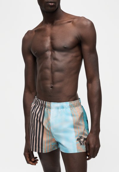 Homme torse nu portant un short de bain court avec des rayures bleues, beiges et noires, logo tête de Méduse sur la jambe gauche, debout devant un fond uni.