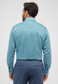 Knoopshirt in teal met geometrische patronen in paars en wit. Lange mouwen en een standaard kraag; gecombineerd met donkere broeken.