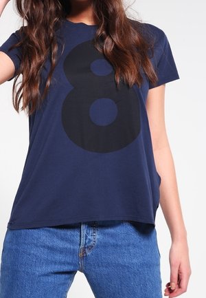 Femme portant un t-shirt bleu marine avec un grand numéro noir 8 et un jean en denim bleu sur un fond uni.