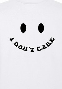 Witte T-shirt met een grote zwarte smileyface met twee ovale ogen en de tekst "IK KAN ME NIETS TROUWEN" in een speelse, gebogen lettertype eronder.