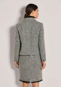 Frau, die mit dem Rücken zur Kamera steht, in einem grauen Tweed-Jackenkostüm mit einem einzelnen Reißverschluss auf dem Rock und einem kleinen Schlitz hinten.