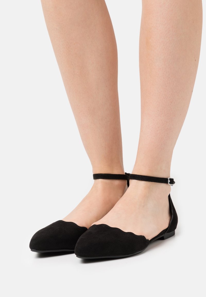 Ballerines en suède noir avec un design festonné, une bride à la cheville, un bout pointu et une semelle plate. La texture est lisse et douce.