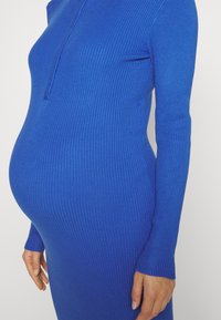 Långärmad, ribbade klänning i livlig blå färg, figurnormande runt midjan, med en halv dragkedja vid halsen och textilstruktur.