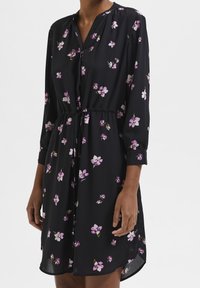 Vestido negro hecho de una tela ligera con cuello, mangas tres cuartos y un estampado floral en púrpura y blanco. Presenta una cintura elástica.