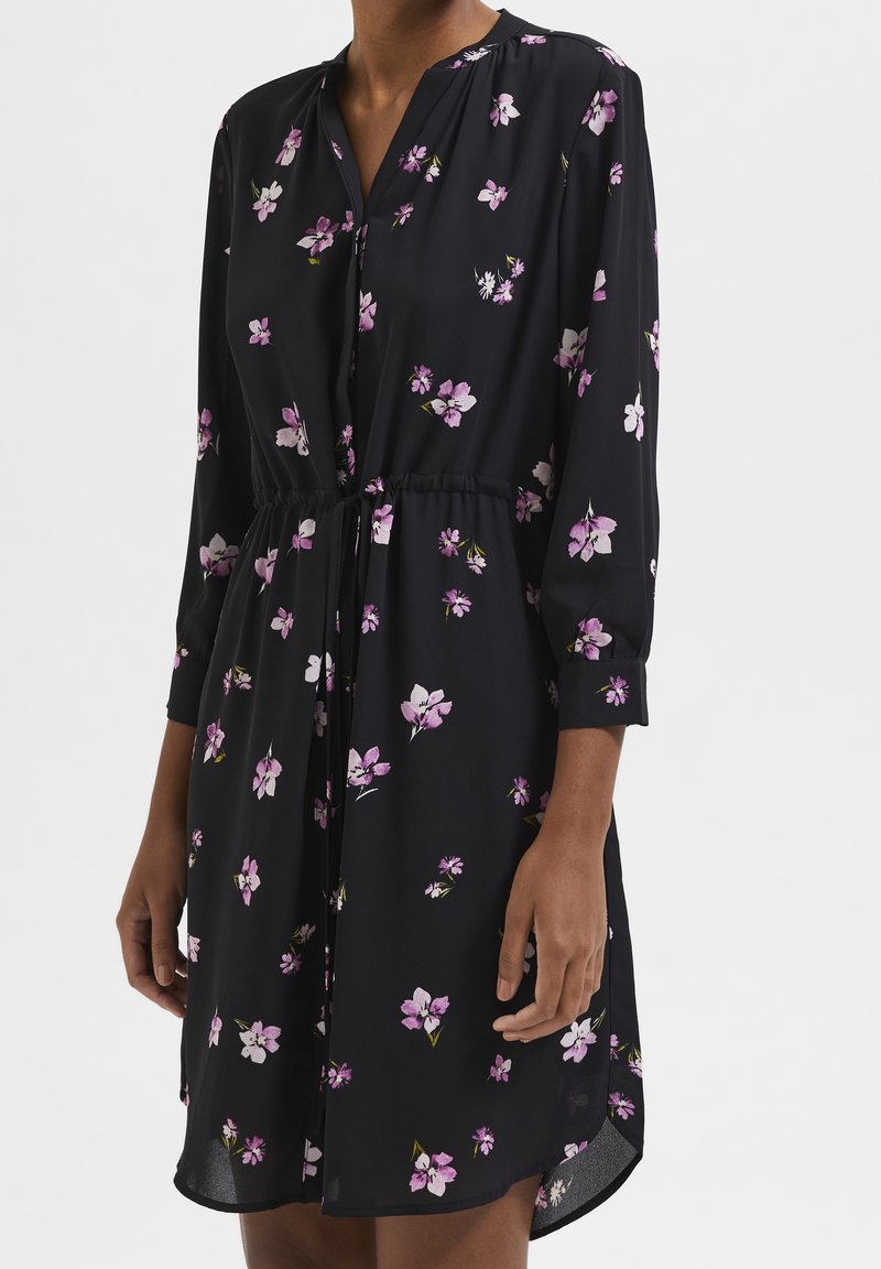 Vestido negro hecho de una tela ligera con cuello, mangas tres cuartos y un estampado floral en púrpura y blanco. Presenta una cintura elástica.