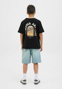 Camiseta de algodón negra con un estampado gráfico que presenta una forma de arco y un diseño de paisaje en la parte posterior, combinada con pantalones cortos de mezclilla azul claros.