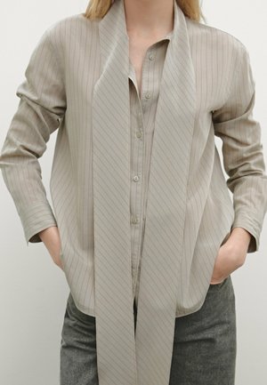 Beżowa koszula w cienkie prążki z guzikami, z długim dopasowanym szalem, noszona z ciemnoszarymi spodniami, ręce włożone do kieszeni.