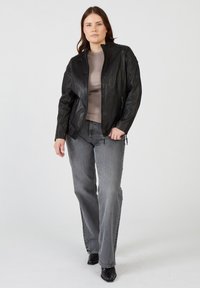 Schwarze Lederjacke mit Stehkragen, Reißverschlussfront und strukturierten Schultern. Getragen über einem beigen Oberteil und grauen, schmal geschnittenen Jeans mit schwarzen Stiefeln.