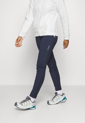 Osoba ubrana w biały polar, granatowe obcisłe spodnie z kieszenią na zamek oraz białe, szaro-turkusowe buty trekkingowe idąca w lewo.