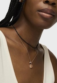 Czarna choker z koralików warstwowy z delikatnym naszyjnikiem w różowym złocie, który ma okrągły wisiorek o charakterystycznym wzorze. Skóra ma średni brązowy odcień.