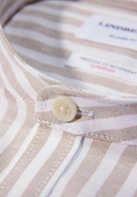 Colletto di camicia a righe marrone chiaro e bianche con dettaglio a bottone, tessuto testurizzato diagonale e etichette per materiali e movimento.