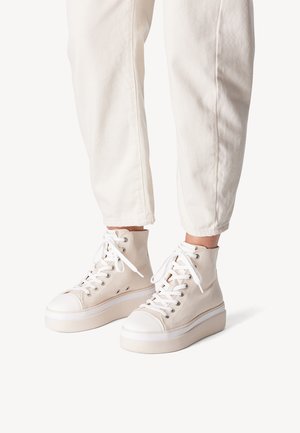Person wearing white high-top platform sneakers with white laces and off-white pants cropped above the ankles against a neutral background.