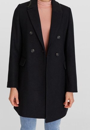 Femme portant un manteau noir croisé sur un col roulé rose et un jean bleu clair, debout devant un fond uni.